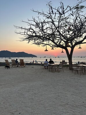 Ein Sitzbereich am Strand mit Stühlen und Tischen, einem Baum und Blick auf das Meer mit Booten.