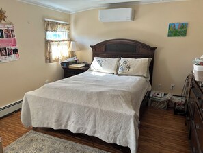 A bedroom with a bed, a nightstand, a lamp, a dresser, a window with curtains, and a wall-mounted air conditioner.
