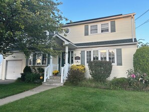 A two-story house with a front porch, a garage, and a well-maintained lawn. - Fabulous 4-bedroom house in East Meadow perfect for your getaway (East Meadow)