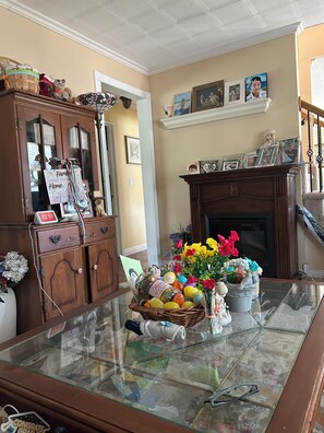 A room with a glass-top table, wooden cabinets, a fireplace, and a wall-mounted shelf with framed photos. - Fabulous 4-bedroom house in East Meadow perfect for your getaway (East Meadow)