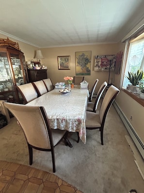 A dining room with a large table, six chairs, a cabinet with glass doors, and a window with plants. - Fabulous 4-bedroom house in East Meadow perfect for your getaway (East Meadow)