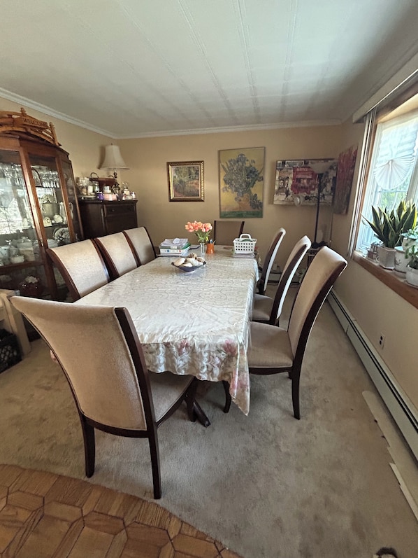 A dining room with a large table, six chairs, a cabinet with glass doors, and a window with plants.