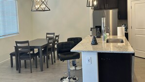 Ett modernt kök med en central köksö, barstolar, ett matbord med stolar, hängande lampor, ett kylskåp och ett fönster med persienner.