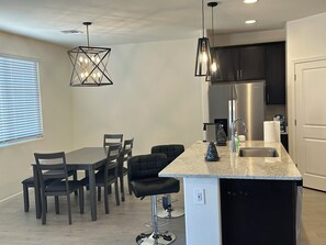 A modern kitchen with a central island, bar stools, a dining table with chairs, pendant lights, a refrigerator, and a window with blinds. -  4-bedroom in superb Tucson, Fully Furnished Escape Near Scenic Desert Trails (Tucson)