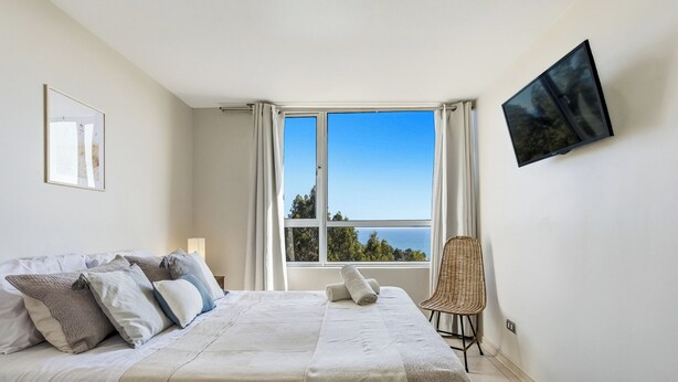 A bedroom with a bed, a chair, a window with curtains, and a wall-mounted TV. - Relaxing Family Condo with Ocean View & Pools in Costa Algarrobo (El Yeco)