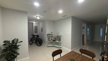 A dining area with a wooden table, chairs, a potted plant, a glass cabinet with bottles, and bicycles in the background.
