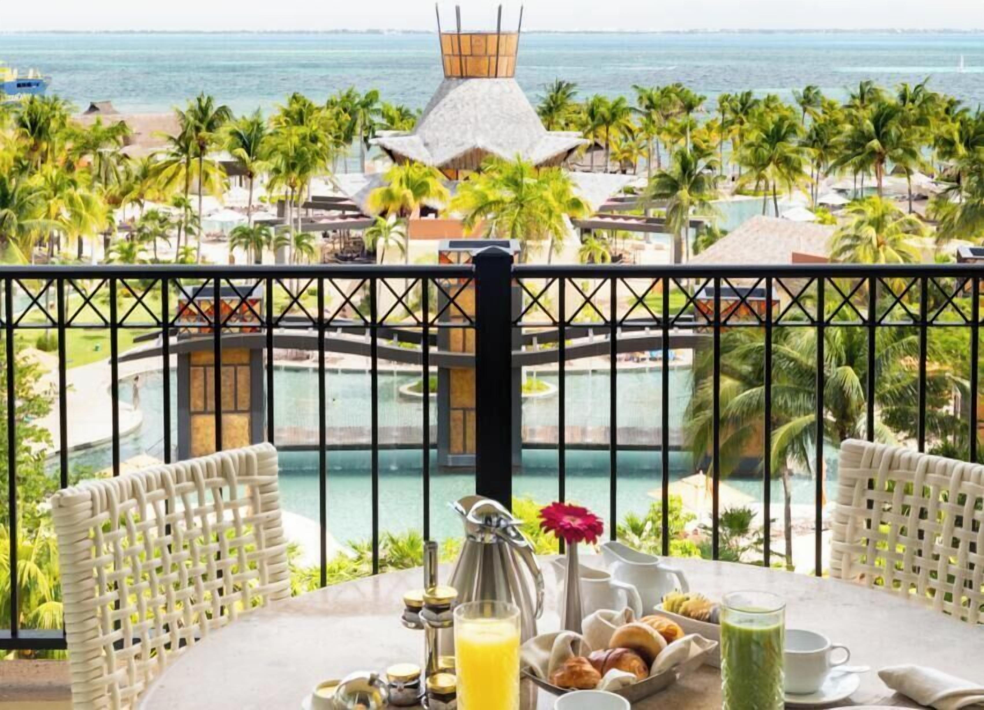 Un balcón con mesa lista para el desayuno con vista a un complejo turístico con piscina y palmeras.