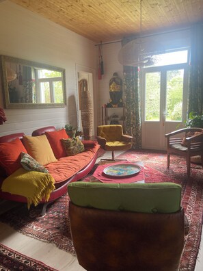 A living room with a red sofa, yellow armchair, and a green ottoman. There is a large mirror on the wall, a Buddha statue, and a window with curtains. - Typical single-storey Médoc house with outbuildings and swimming pool (Carcans)