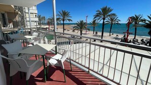 Un balcone con sedie e tavoli di plastica bianca, affacciato su una spiaggia con palme e un cielo azzurro terso.
