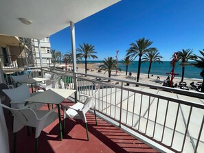 Ein Balkon mit weißen Plastikstühlen und -tischen, der Blick auf einen Strand mit Palmen und einem klaren, blauen Himmel gewährt.