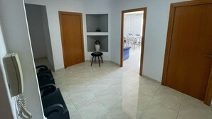 Un corridoio con pavimento piastrellato, porte in legno, un tavolino con un vaso e una panca blu.