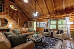 A log cabin interior with a staircase, leather sofas, a coffee table, and a large window with a view. - Private Cabin in Gatlinburg | Hot Tub + Games + Fire Pit + Scenic Decks (Gatlinburg)