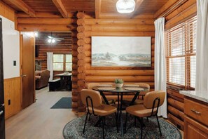A log cabin interior with a dining area featuring a round table and chairs, a painting on the wall, and a view through a window with blinds. - Private Cabin in Gatlinburg | Hot Tub + Games + Fire Pit + Scenic Decks (Gatlinburg)