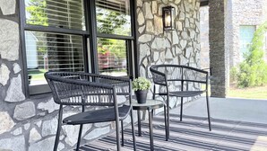 Un mur extérieur en pierre avec une fenêtre, deux chaises en osier, une petite table avec un pot de fleurs et un pilier en pierre.