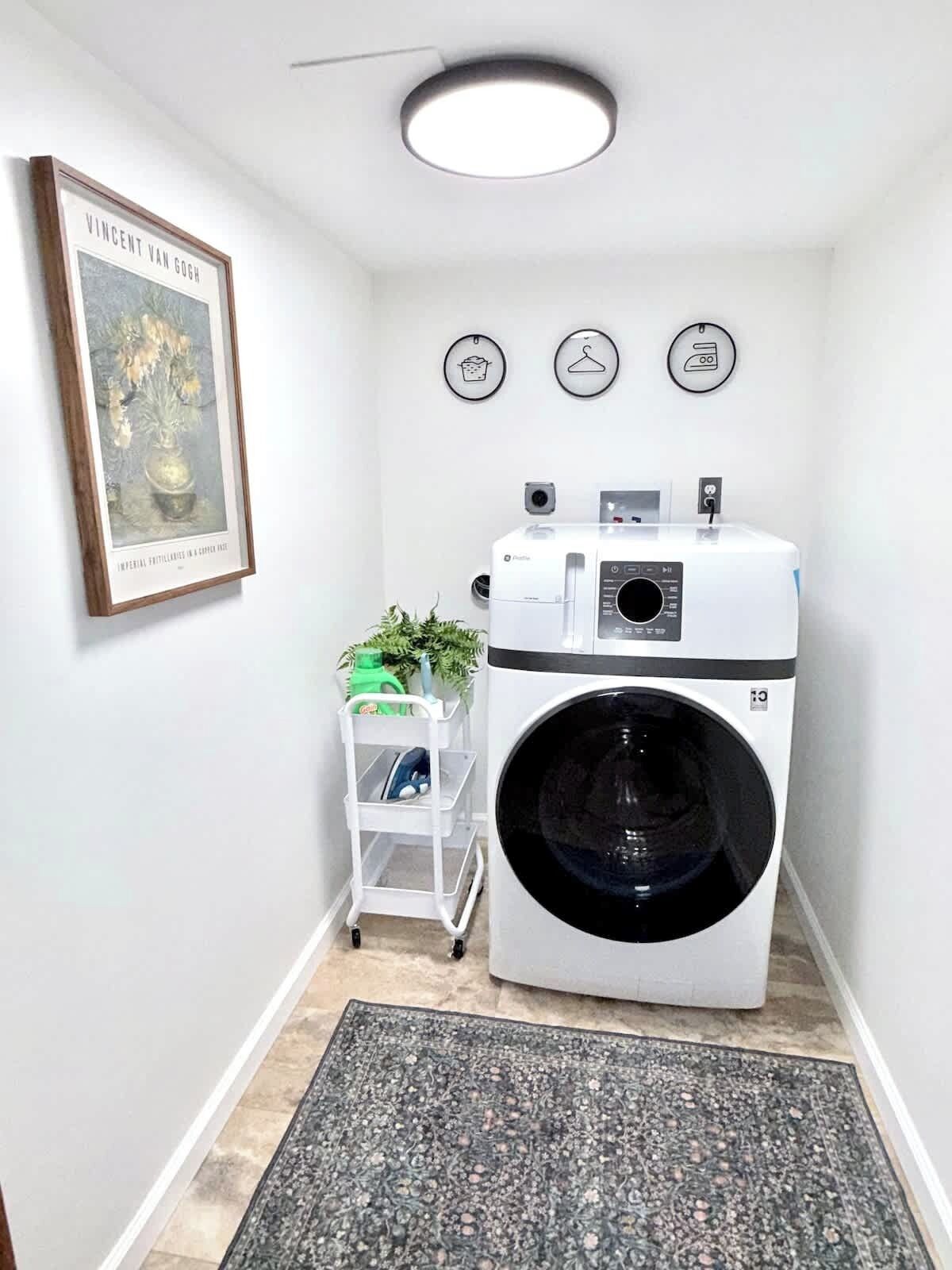 Un espace buanderie comprenant un lave-linge et un sèche-linge à chargement frontal, un petit chariot contenant des produits de nettoyage, une œuvre d’art encadrée et un luminaire au plafond.