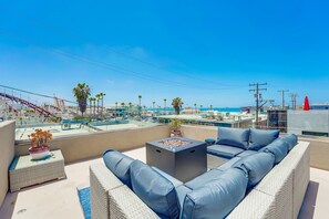Eine Dachterrasse mit einer Couch, einem kleinen Tisch und einem Blumentopf. Von der Terrasse hat man Blick auf einen Strand und eine Stadtlandschaft.