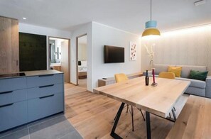 A modern kitchen with a built-in stove, a dining area with a wooden table and chairs, and a living area with a sofa and a TV mounted on the wall.