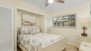 Une chambre à coucher avec un lit, une fenêtre avec des stores, un ventilateur de plafond et une table de chevet avec une lampe.