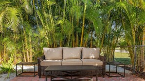 Un canapé beige agrémenté de coussins, une table basse en bois et deux petites tables d’appoint, le tout disposé sur une terrasse pavée, avec des palmiers en arrière-plan.