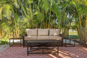 A beige sofa with cushions, a wooden coffee table, and two small side tables, all set on a paved patio with palm trees in the background.