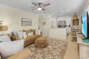 A living room with a sofa, a round coffee table, and a TV. There is a kitchen area with a refrigerator and a dining table.