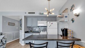 A kitchen with grey cabinets, a white microwave, a black refrigerator, and a coffee maker. There are two bar stools and a chandelier. - Ocean Forest Plaza 912 (Myrtle Beach)