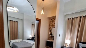 Ein Hotelzimmer mit einem großen Spiegel, einem Bett, einem Nachttisch mit Lampe, einem kleinen Badezimmer und einem gewebten Korb mit Handtüchern.