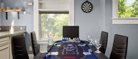 A poker table with chips and cards, a dartboard, and a window with a view of trees.