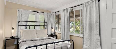A bedroom with a black metal bed frame, two bedside tables with lamps, and white curtains on the windows.