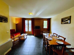A dining area with a table set for four, a sofa, a TV, and a window with curtains. - 1st floor, swimming pool, sauna, balcony, parking, tv, ski locker, 49m², Saint Gervais (Saint-Gervais-les-Bains)