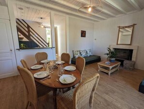 A dining area with a wooden table and wicker chairs, a staircase, a sofa, a fireplace, and a mirror. - Gîte Bambou - Country charm and heated swimming pool in the heart of Charente (Chadurie)