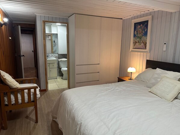 A bedroom with a bed, a nightstand with a lamp, a wooden chair, a wardrobe, and a bathroom visible through an open door.