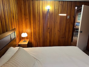 Habitación con paneles de madera, una cama, una mesita de noche con lámpara y una puerta que da a otra habitación.