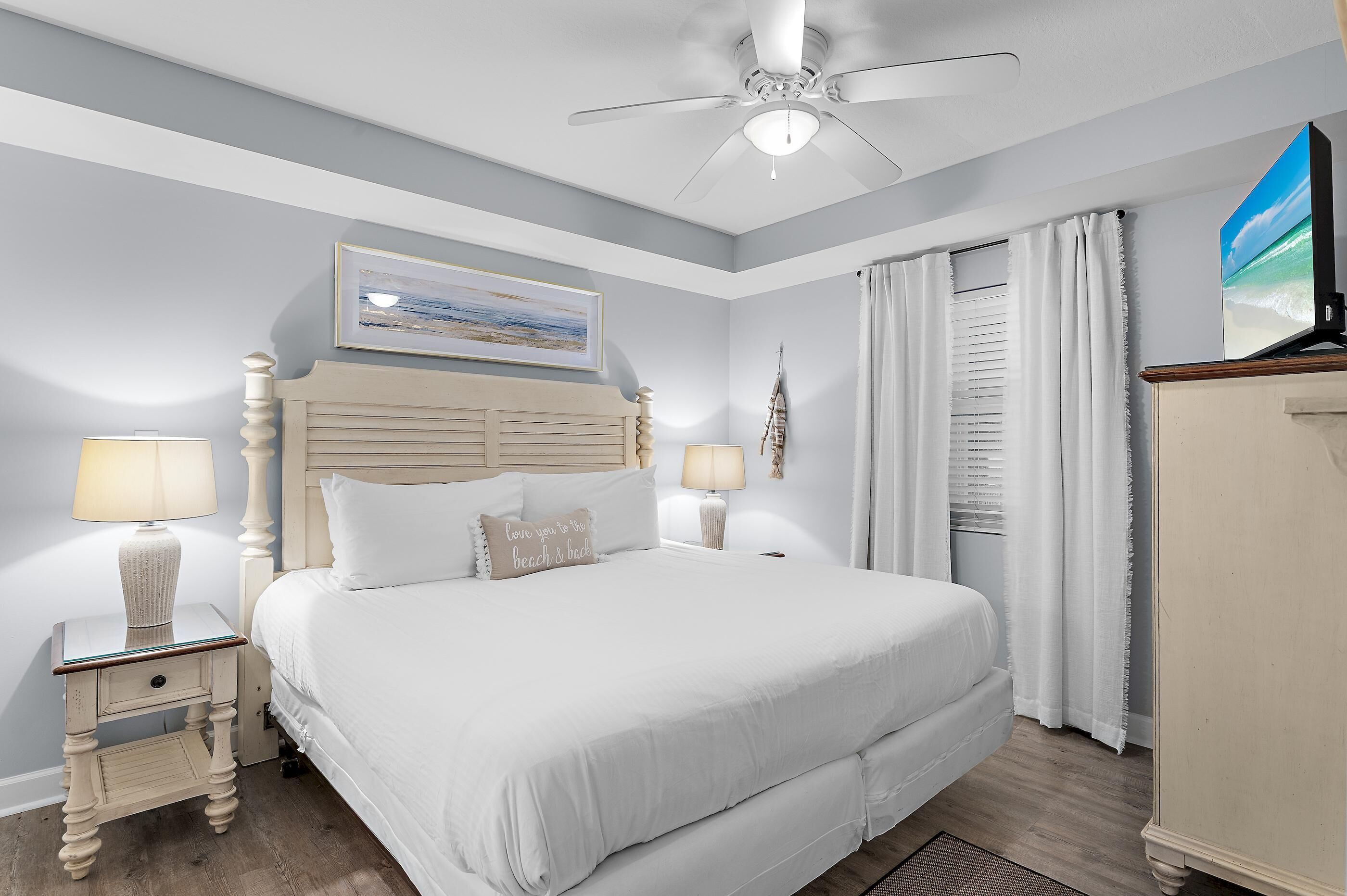 Un dormitorio ordenado con cama, lámparas de noche, ventilador de techo, un cuadro y una ventana con cortinas.