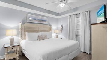 Un dormitorio ordenado con cama, lámparas de noche, ventilador de techo, un cuadro y una ventana con cortinas.