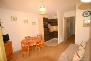 Ein Wohnzimmer mit einer Couch, einem Esstisch mit Stühlen, einem Fernseher und einer Küche im Hintergrund.