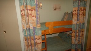 Ein Stockbett mit Holzbettgestell und einer grünen Matratze, Vorhänge mit geometrischem Muster sowie eine kleine Wandleuchte.