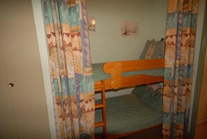 Ein Stockbett mit Holzbettgestell und einer grünen Matratze, Vorhänge mit geometrischem Muster sowie eine kleine Wandleuchte.