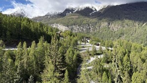 Un paysage montagneux avec une forêt dense, un bâtiment doté d’un toit en pente et, au loin, des sommets enneigés.
