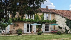 A rustic stone building with a tiled roof, white shutters, and a garden with a lawn and outdoor seating.