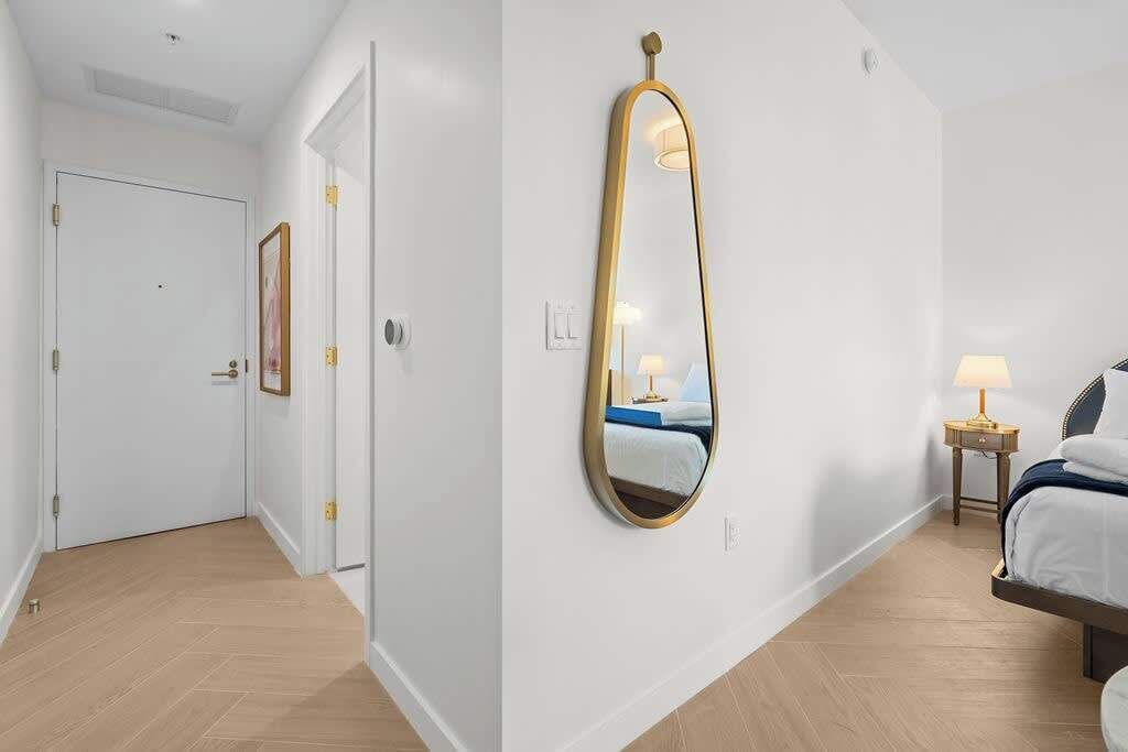 A hotel room with a large mirror, a bed, a bedside table with a lamp, and a door.