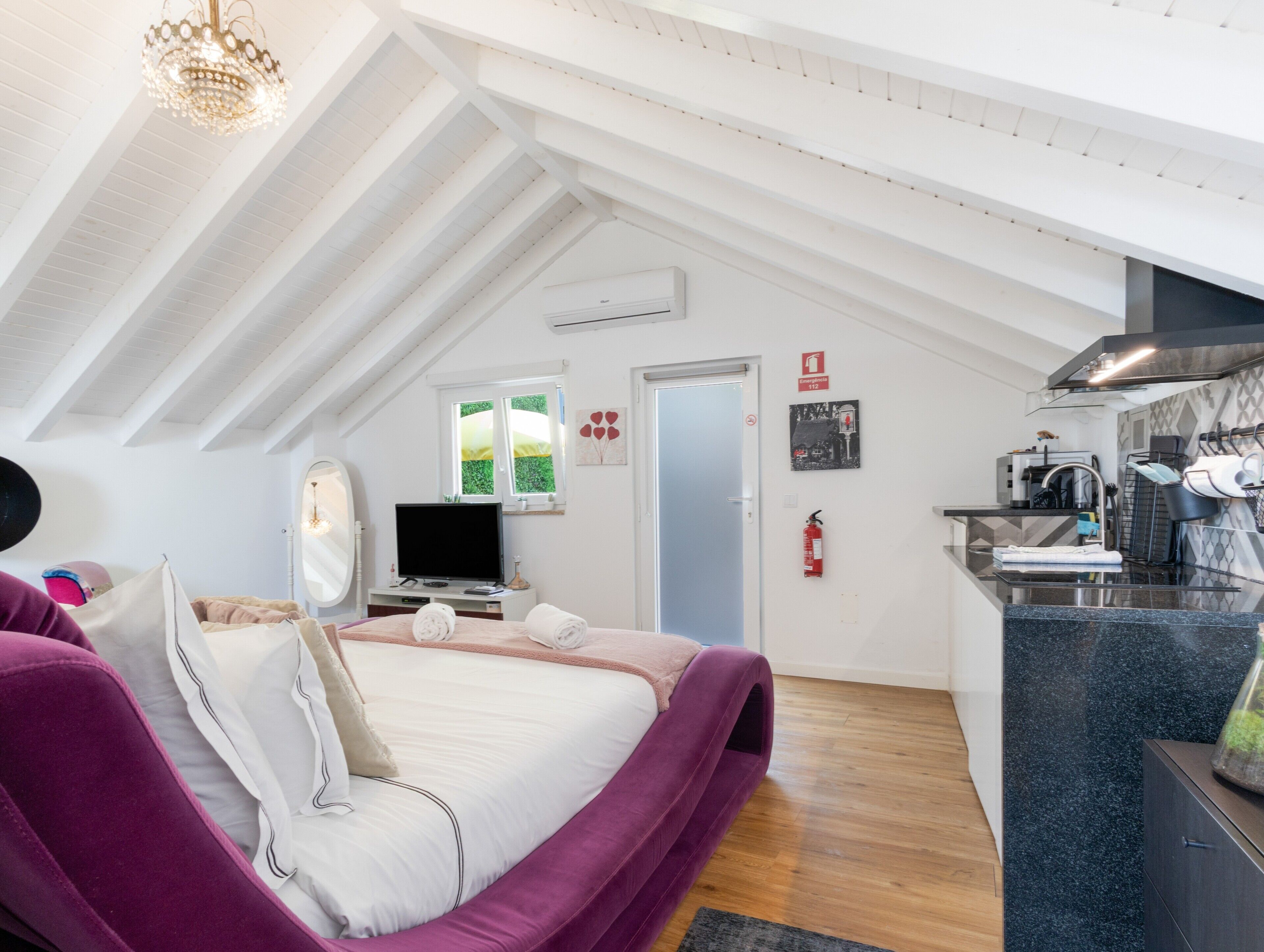 A bedroom with a bed, a TV, a mirror, a window, and a kitchenette.