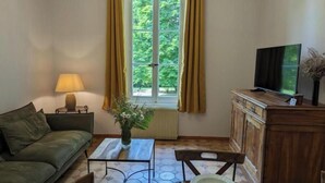 Una habitación con sofá, una mesita con lámpara, una mesa de comedor para dos personas, televisor y ventana con cortinas.