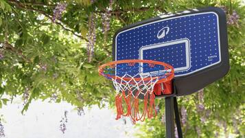 Un aro de baloncesto con red, sujeto a un tablero con un logotipo, sobre un fondo de follaje verde y flores púrpuras.