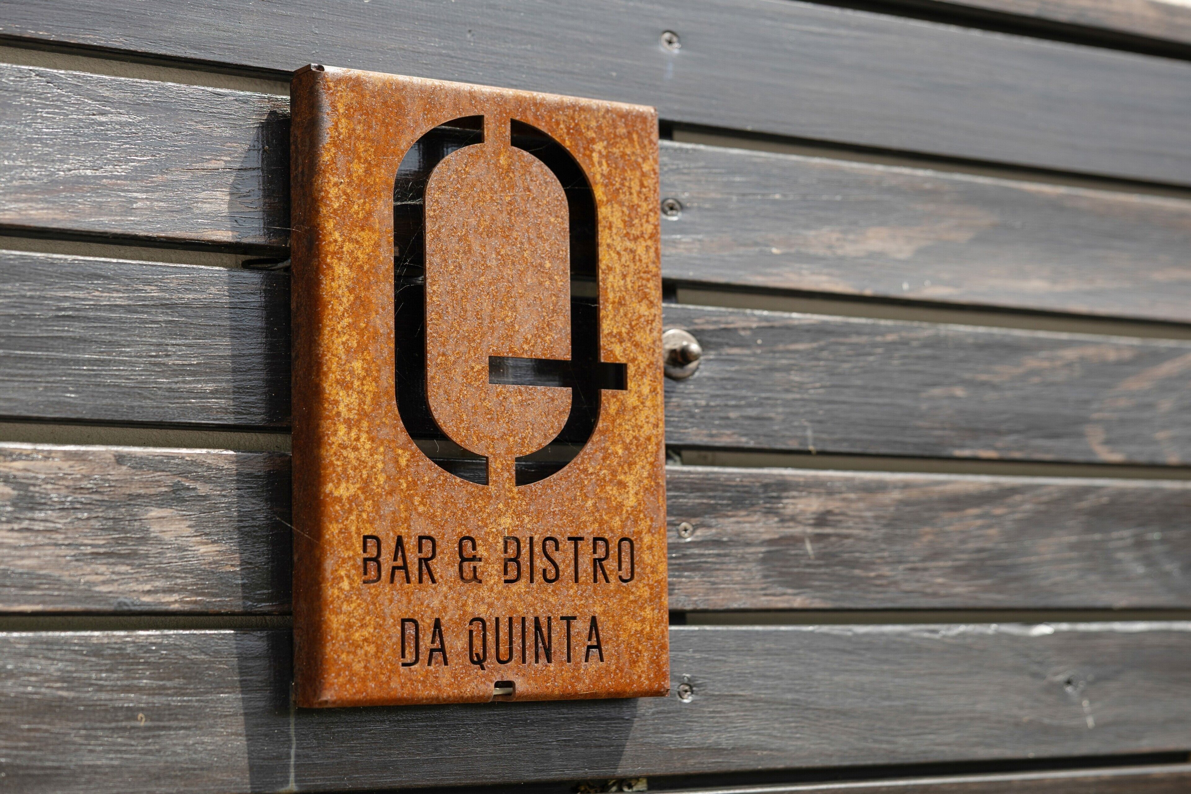 Een metalen bord met een logo en de tekst 'BAR & BISTRO DA QUINTA' bevestigd aan een houten wand.