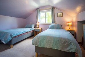 A bedroom with two single beds, a window with curtains, bedside lamps, and a framed picture on the wall. - Oaks Lodge Cottage (Ambleside)