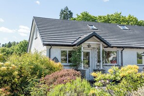 A house with a sloped roof, a porch, and a garden with various plants. - Oaks Lodge Cottage (Ambleside)