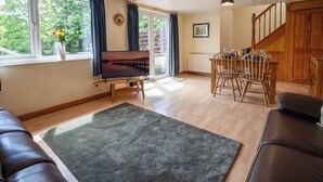 Sala de estar com sofá, televisão, mesa de jantar com cadeiras, escada e vista para árvores pela janela.