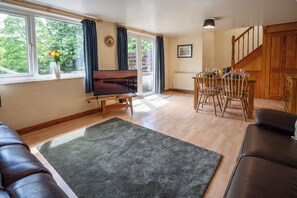 A living room with a sofa, a TV, a dining table with chairs, a staircase, and a view of trees through the window. - Oaks Lodge Cottage (Ambleside)