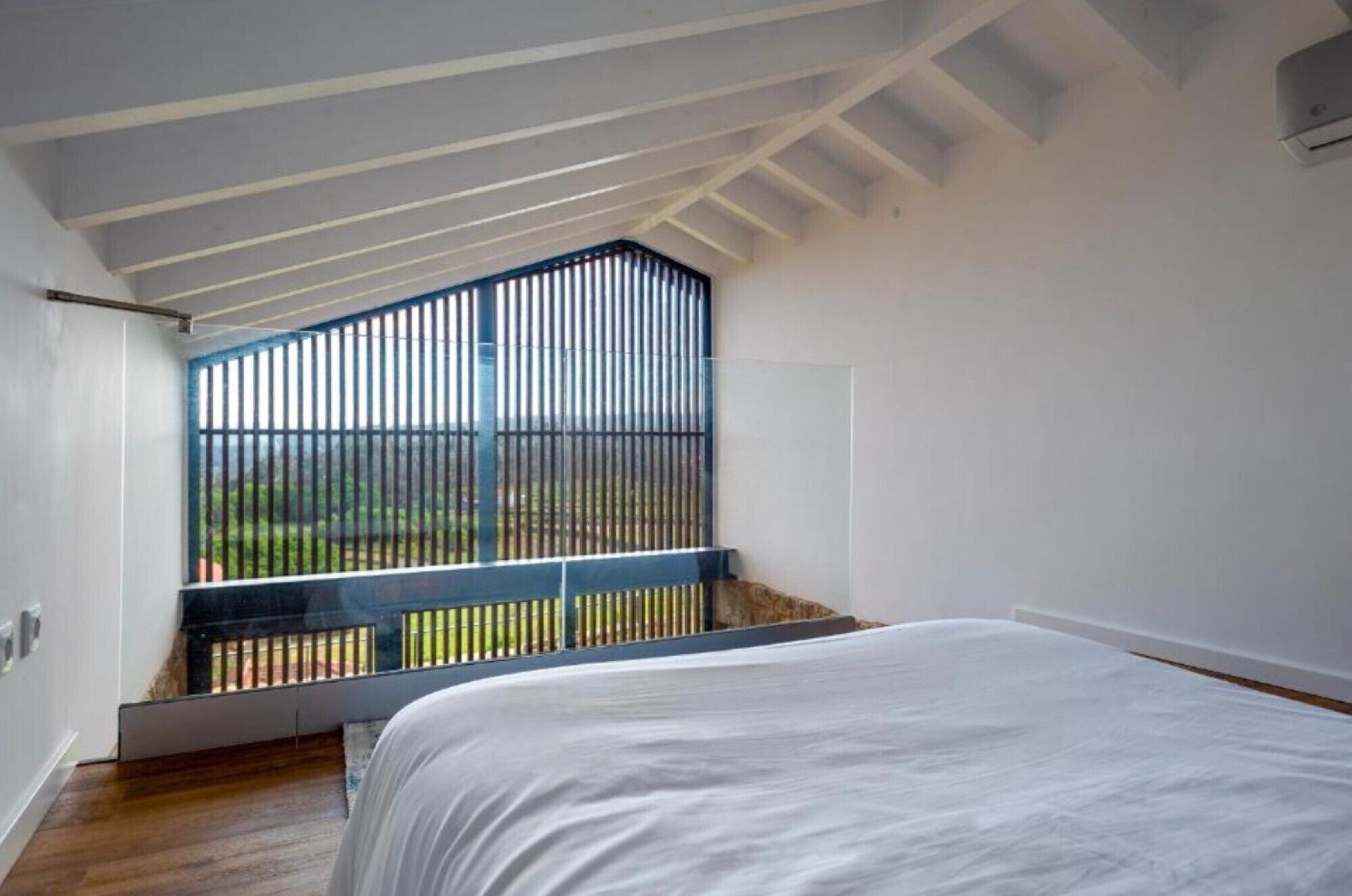 A bedroom with a sloped ceiling, a large window with vertical blinds, a bed with white bedding, and a view of greenery outside.